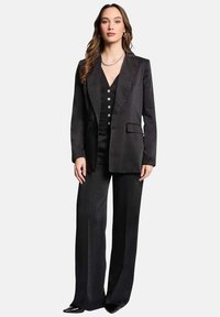 Traje negro a medida que presenta un blazer entallado con solapas de muesca y pantalones de pierna ancha. El material tiene una textura suave con un ligero brillo.
