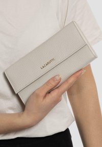 Billetera de cuero texturizado gris claro con solapa plegable y logo dorado, sostenida en una mano contra un fondo de camiseta blanca lisa.