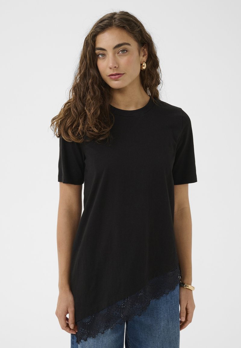 Tânără cu păr lung și ondulat, purtând un top negru asimetric cu mâneci scurte și dantelă, și blugi albaștri, stând în fața unui fundal simplu.