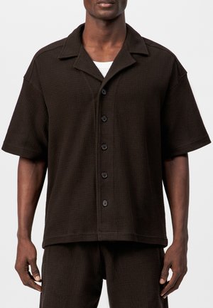 Mann trägt ein dunkelbraunes kurzärmeliges Hemd mit Knöpfen und passende strukturierte Shorts, mit sichtbarem weißen Kragen des Unterhemds.