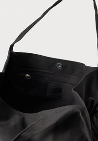 Čierna taška na plátno s vnútorným vreckom na zips, zapínaním na cvok a dvojitými textilnými popruhmi. Vo vnútri má nenápadnú štítkovú značku.