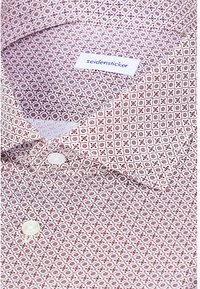 Chemise blanche avec petit motif géométrique rouge, boutons blancs et étiquette de col portant la mention "seidensticker" en texte bleu.