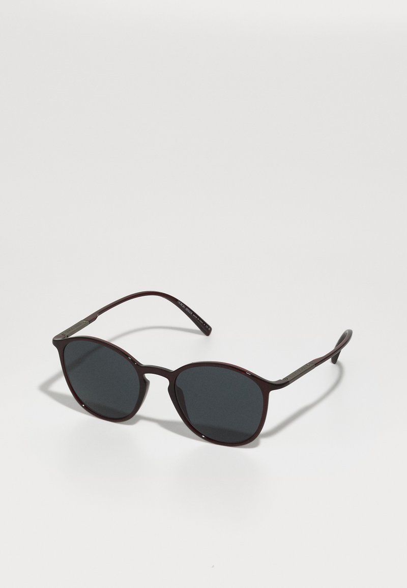 Burgundy sunglasses with dark lenses, rounded shape, glossy finish, and slender arms featuring metallic accents and engraved details.