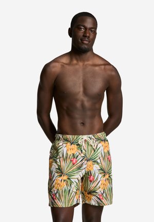 Homme musclé portant un short de bain tropical à fleurs avec des feuilles vertes et des fleurs jaunes et rouges, debout avec les mains derrière le dos.