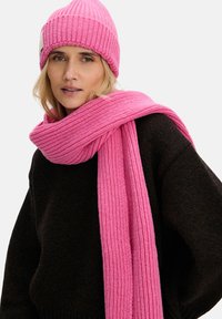 Różowa prążkowana dzianinowa czapka i pasujący szalik, noszone z ciemnobrązowym swetrem. Szalik luźno owinięty wokół szyi, ukazujący dzianinową fakturę.