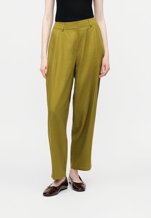 Γυναίκα που φοράει χακί παντελόνια με φαρδιά μπατζάκια και ψηλή μέση, καθώς και σκούρα καφέ μπαλαρίνες, στέκεται μπροστά σε μια απλή φόντο.