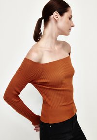 Top acanalado, con los hombros al descubierto en color naranja quemado, con mangas largas ajustadas y una silueta ceñida, combinado con pantalones oscuros.