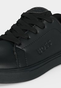 Gros plan d'une chaussure de sport en cuir noir avec des lacets noirs et le mot BOSS embossé sur le côté.