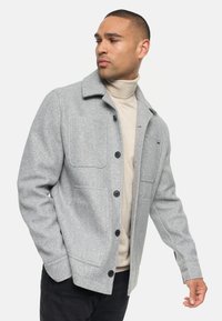 Veste en mélange de laine grise avec un col, quatrepoches avant et boutons noirs. Portée sur un col roulé beige, offrant une coupe décontractée et une texture lisse.