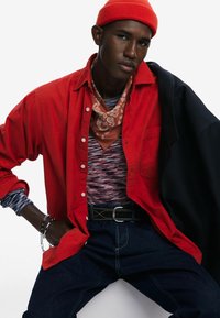 Punane särk nööpidega triibulise pika varruka topi peal, kombineeritud pruuni bandana ja tumedate teksadega. Must jakk on visatud ühe õla peale.