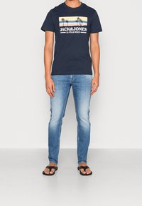 Navy blue cotton t-shirt with palm tree graphic and "JACK&JONES LA JOLLA BEACH" text above, paired with light blue jeans and black flip-flops.
