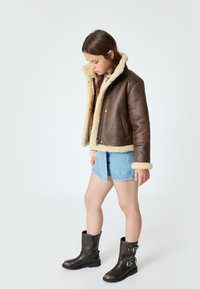 Braune Lederjacke mit cremefarbenem Kunstfellfutter, Reißverschluss und Druckknöpfen. Tragen kombiniert mit Jeansshorts und schwarzen Stiefeletten.