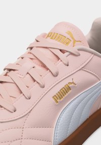 Κοντινό πλάνο ροζ αθλητικού παπουτσιού Puma με ανοιχτό μπλε λωρίδα στην πλευρά, χρυσά λογότυπα Puma στη γλώσσα και την πλευρά, και ροζ κορδόνια δεμένα σε φιόγκο.