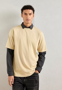 Bežinis trumpomis rankovėmis marškinėliai, uždėti ant juodų dryžuotų ilgomis rankovėmis marškinėlių. Modelis dėvi tamsias džinsines kelnes, rankas kišenėse. Lygus paviršius.