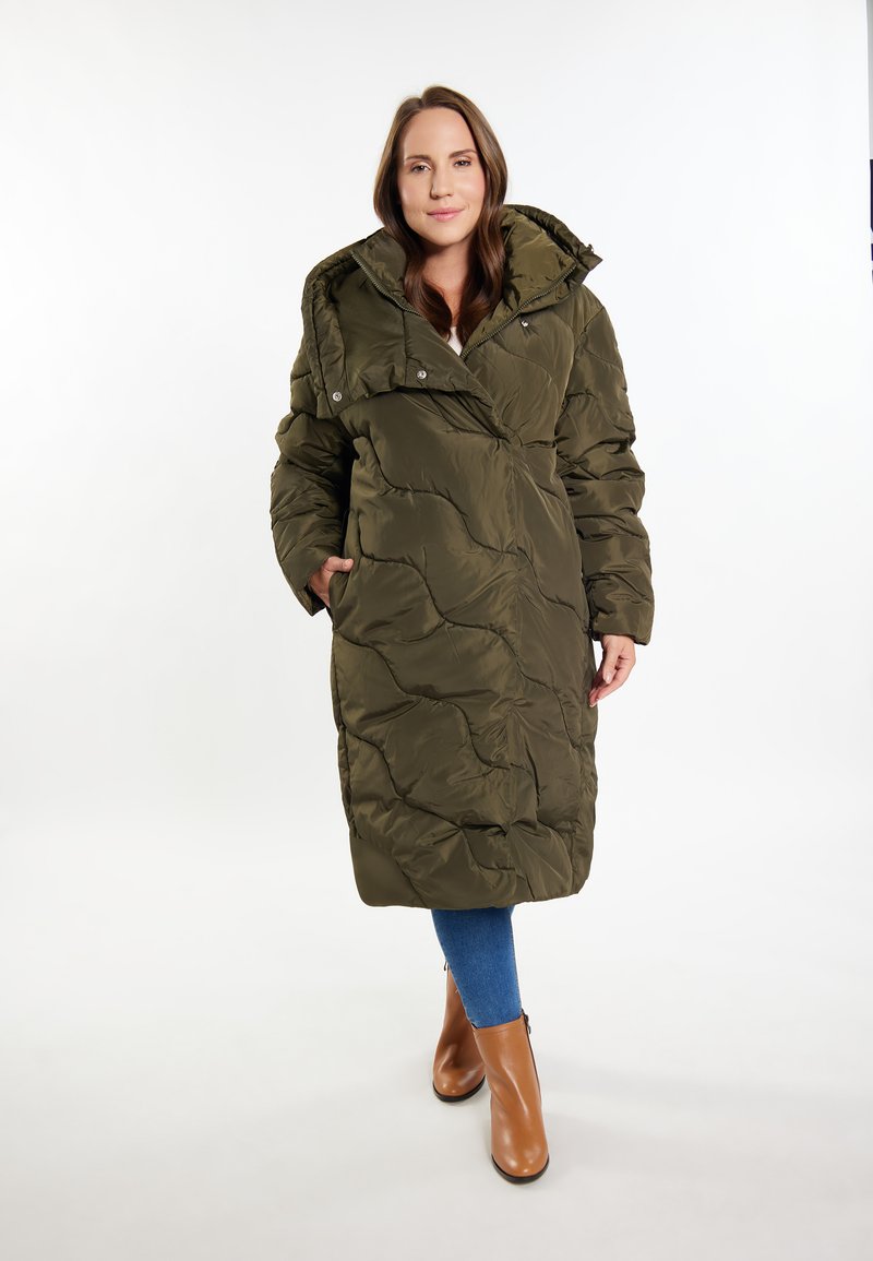 Lang olivengrøn puffjakke med en quiltet design, stor krave og knaplukninger. Sammen med blå jeans og brune ankelstøvler.