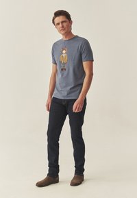 Modré bavlněné tričko s vyšitým motivem medvěda v hnědém svetru, nošené s tmavými džínami a hnědými kotníkovými botami. Jednoduché pozadí.