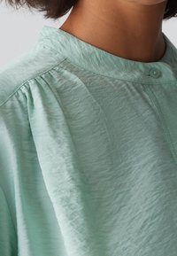 Blusa verde menta in tessuto di trama, con colletto tondo e chiusura a bottoni. Le pieghe alle spalle aggiungono dettaglio.