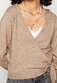 Maglione avvolgente beige in maglia con chiusura a cravatta, design a scollo a V e texture morbida. Le collane sovrapposte includono perle e un pendente a cuore verde.