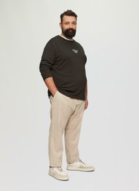 Dunkelbluse mit langen Ärmeln und weißen Akzenten, beigen Cordhosen und hellen Turnschuhen. Die Bluse hat ein kleines Grafikdesign auf der Brust.