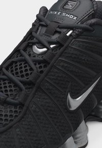 Primer plano de una zapatilla Nike Shox negra que muestra la parte superior de malla, cordones negros, el logo Swoosh plateado y una suela texturizada con amortiguación.