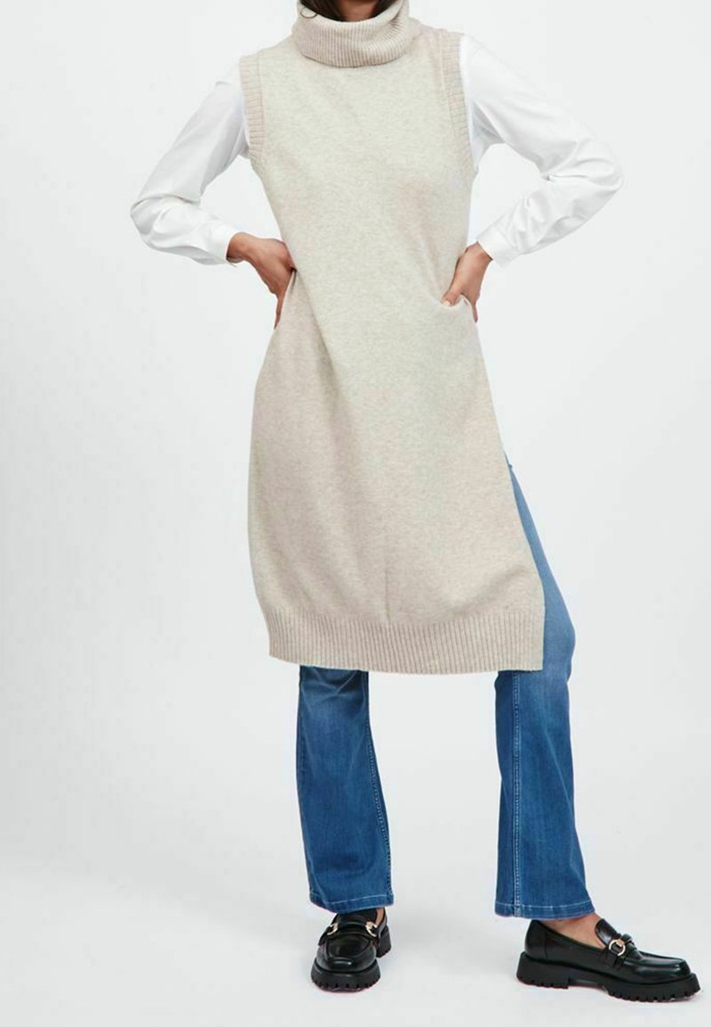 Robe-pull beige clair sans manches à col roulé avec bords côtelés, associée à un jean évasé bleu et des chaussures noires. Texture tricote douce.