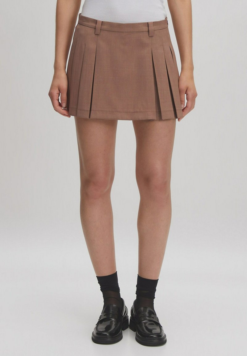 Bruine geplooide rok met een aansluitende taille, gemaakt van textuurstof, gecombineerd met zwarte loafers en gestreepte zwarte sokken.