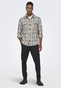 Plaid flannel shirt in light green, lavender, and white, with two chest pockets, buttoned cuffs, and a relaxed fit, paired with black cargo pants.