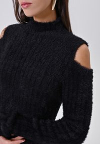 Maglione nero in maglia fuzzy con collo alto e spalle scoperte. Il tessuto ha una consistenza morbida con un design a coste.