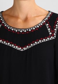 Blouse noire avec un col rond orné de broderies florales rouges et blanches le long du col et des épaules. Texture de tissu lisse.