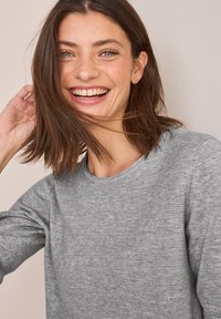 Vaaleanharmaa teksturoitu neulepaita, jossa on pyöreä pääntie ja pitkät hihat, jota käyttää henkilö, jolla on keskikokoa oleva ruskea hius, ja joka hymyilee neutraalia taustaa vasten.