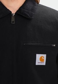 Černá bunda s manžetovým límcem, předním zipem a hrudní kapsou, která má obdélníkovou nášivku s oranžovým logem na bílé látce.