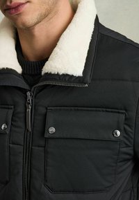 Jachetă neagră căptuşită, cu un guler de blană sintetică albă, închidere cu fermoar și două buzunare cu clapetă și nasturi. Material texturat cu model matlasat.