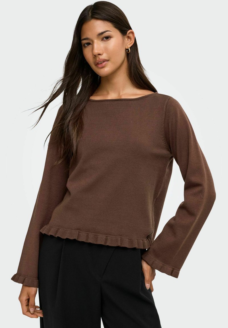 Jeune femme aux longs cheveux foncés portant un haut marron à manches longues avec des bords volantés et un pantalon noir, debout devant un fond uni.