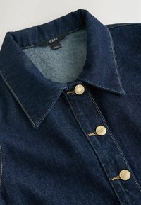Col de veste en denim bleu foncé avec trois boutons en métal et coutures visibles, étiquetée "NEXT" et fabriquée au Maroc.