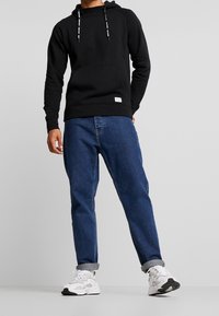 Sudadera negra con capucha y cordones, bolsillo canguro; combinada con jeans de denim azul sueltos y zapatillas blancas con acentos oscuros.