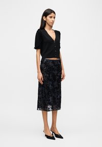 Μαύρη κοντή ζακέτα με κοντά μανίκια με βολάν και κουμπιά, συνδυασμένη με σκούρα midi φούστα με κεντήματα, παγιέτες και διάφανο επικάλυμμα.