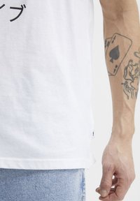 Camiseta blanca de algodón con texto gráfico negro, de corte recto y una pequeña etiqueta lateral. Combinada con shorts de mezclilla claros.