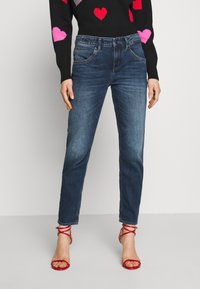 Jeans de color azul oscuro con ajuste slim, que presentan un clásico diseño de cinco bolsillos, cintura de altura media y un ligero desgastado. Combinados con sandalias rojas de tiras.