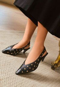 Fekete, arany szegecsekkel díszített slingback kisméretű sarkú cipőt viselő nő, bézs textúrájú szőnyegen ül, fa padlón.