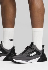 Zapatillas deportivas grises con detalles en negro y blanco. Parte superior de malla, suela gruesa, marca prominente. Llevadas con calcetines blancos de canalé.
