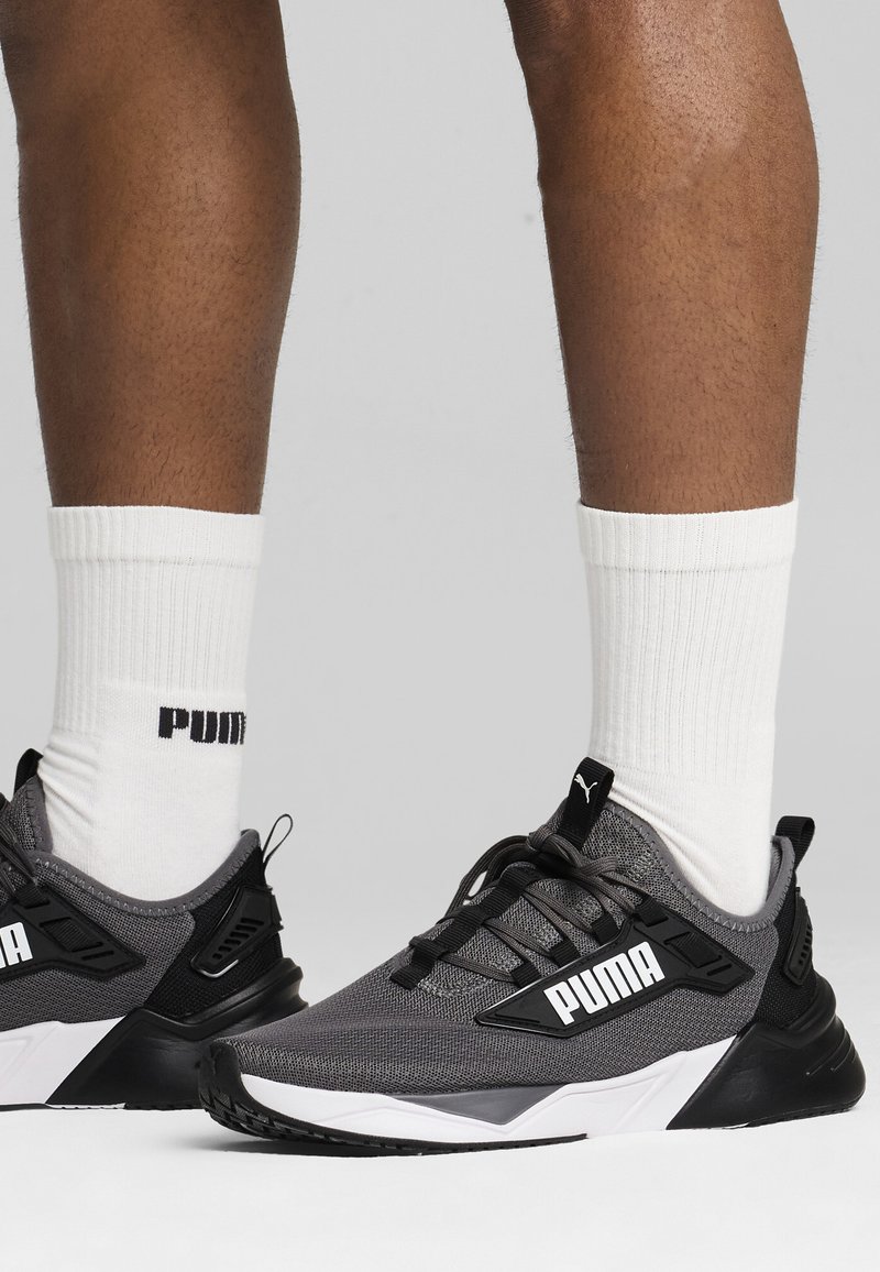 Zapatillas deportivas grises con detalles en negro y blanco. Parte superior de malla, suela gruesa, marca prominente. Llevadas con calcetines blancos de canalé.