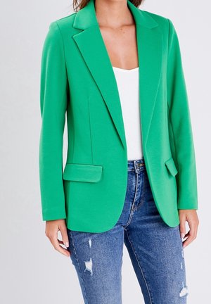 Femme portant un blazer vert vif sur un haut blanc avec un jean bleu déchiré, se tenant contre un fond clair.