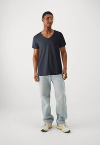 Námořnicky modré tričko s výstřihem do V z měkké látky, v kombinaci s světle modrými džínami a bílými teniskami se žlutými detaily. Model má na sobě brýle se žlutým tónováním.