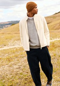 Beige Fleecejacke mit hohem Kragen, grauer Pullover darunter, dunkelblaue Hose und braune gestrickte Mütze. Außenszene mit grasbewachsenem Gelände.