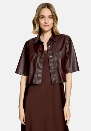 Frau mit blondem Haar, die eine dunkelbraune, kurzärmelige Lederjacke über einem passenden braunen Kleid trägt, steht vor einem schlichten Hintergrund.