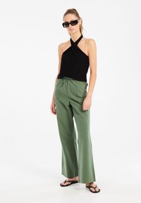 Top negro sin mangas con escote retorcido, combinado con pantalones sueltos de color verde claro y chanclas negras. Modelo usando gafas de sol negras.