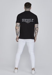 Crna pamučna majica s bijelom "SIKSILK" grafikom na leđima, uparena s bijelim uskim trapericama i bijelim tenisicama, stoji naslonjena na jednostavnu pozadinu.