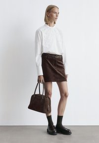 Woman wearing a white embroidered blouse, brown corduroy skirt with belt, black socks, loafers, and carrying a large brown handbag.