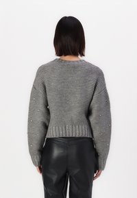 Donna di spalle che indossa un maglione grigio lavorato a maglia con maniche decorate e pantaloni in pelle nera su uno sfondo bianco semplice.