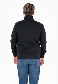 Zwarte ritsvrijheid met een hoge kraag, zachte textuur en lange mouwen, gedragen over blauwe denim jeans. De weergave toont de achterkant van de outfit.